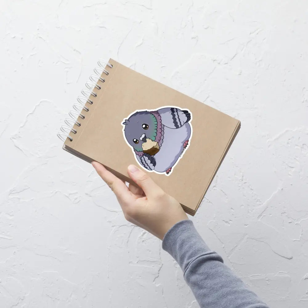 A product mockup of a 5x5" vinyl sticker on a brown notebook cover, featuring Fred the Pigeon.