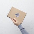 A product mockup of a 3x3" vinyl sticker on a brown notebook cover, featuring Fred the Pigeon.