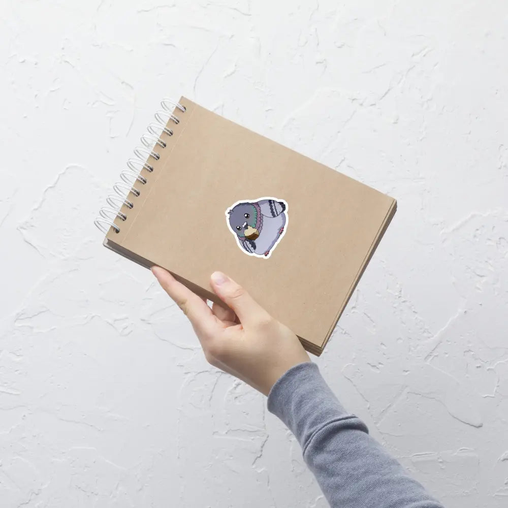 A product mockup of a 3x3" vinyl sticker on a brown notebook cover, featuring Fred the Pigeon.