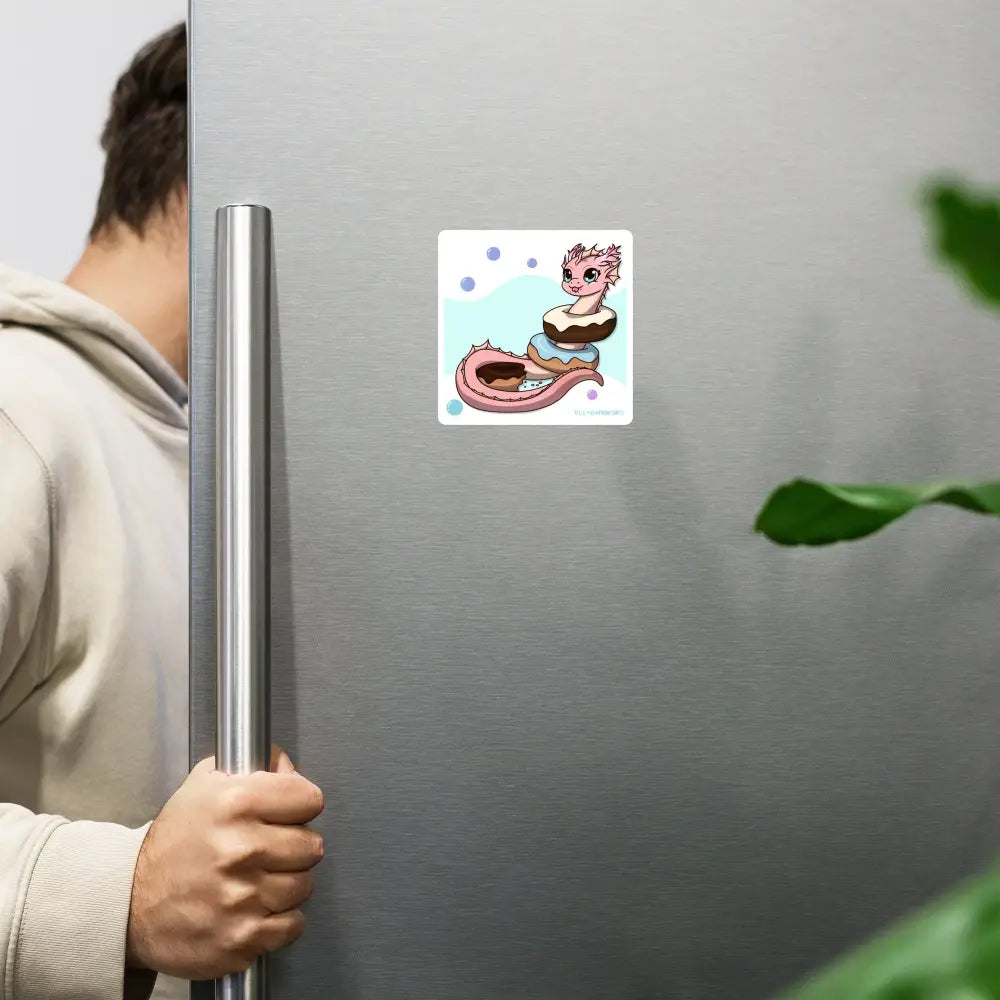 A product mockup of the 4x4" magnet featuring Pearl the Coral Dragon hanging on a stainless steel refrigerator door