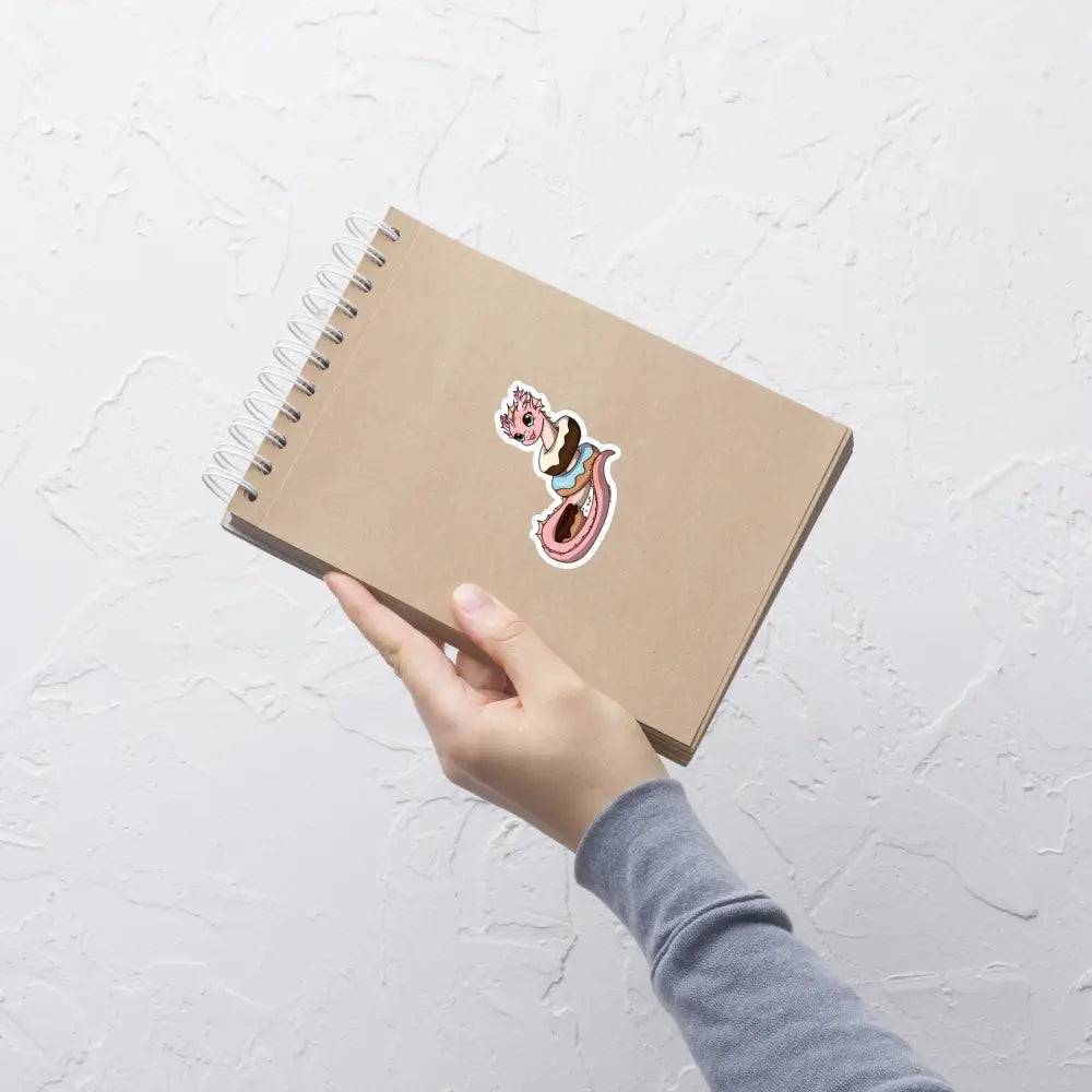 A product mockup of a 3x3" vinyl sticker on a brown notebook cover, featuring Pearl the Coral Dragon.