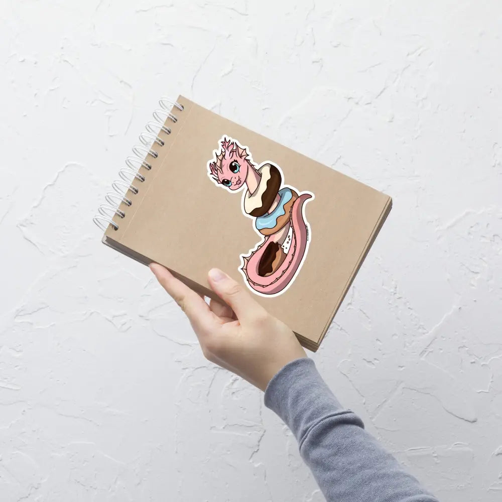 A product mockup of a 5x5" vinyl sticker on a brown notebook cover, featuring Pearl the Coral Dragon.