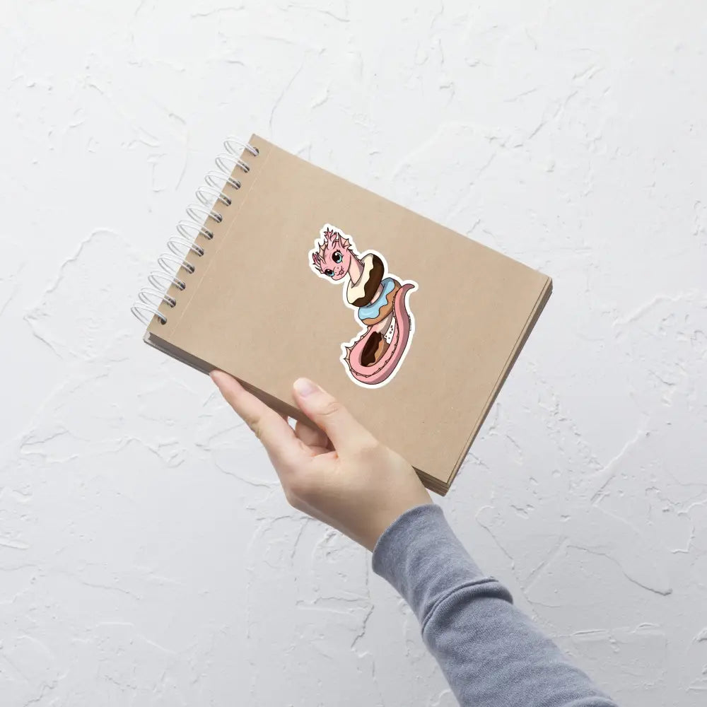 A product mockup of a 4x4" vinyl sticker on a brown notebook cover, featuring Pearl the Coral Dragon.