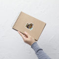 A product mockup of a 3x3" vinyl sticker on a brown notebook cover, featuring Safiya the Sphinx.