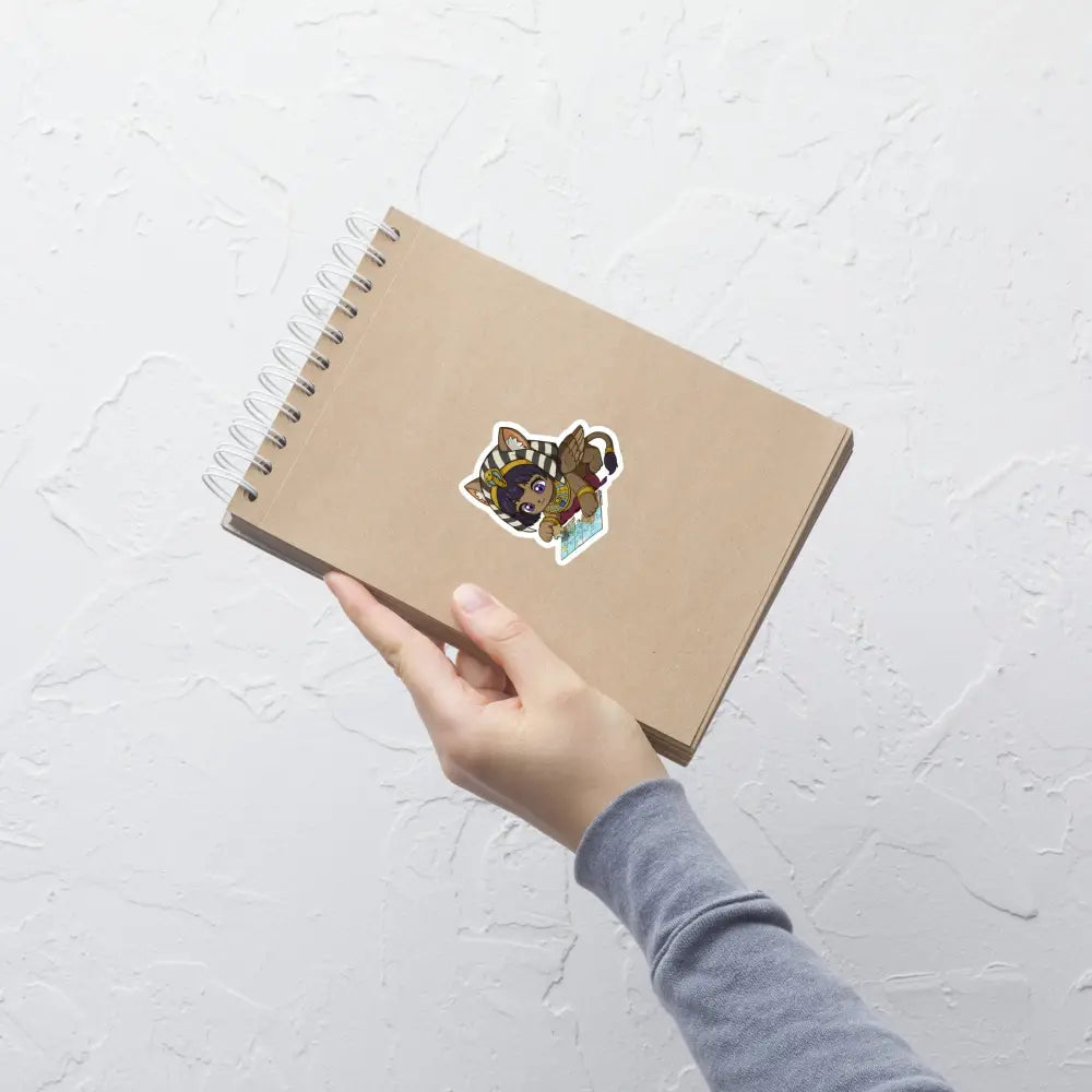 A product mockup of a 3x3" vinyl sticker on a brown notebook cover, featuring Safiya the Sphinx.