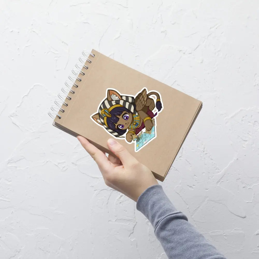 A product mockup of a 5x5" vinyl sticker on a brown notebook cover, featuring Safiya the Sphinx.