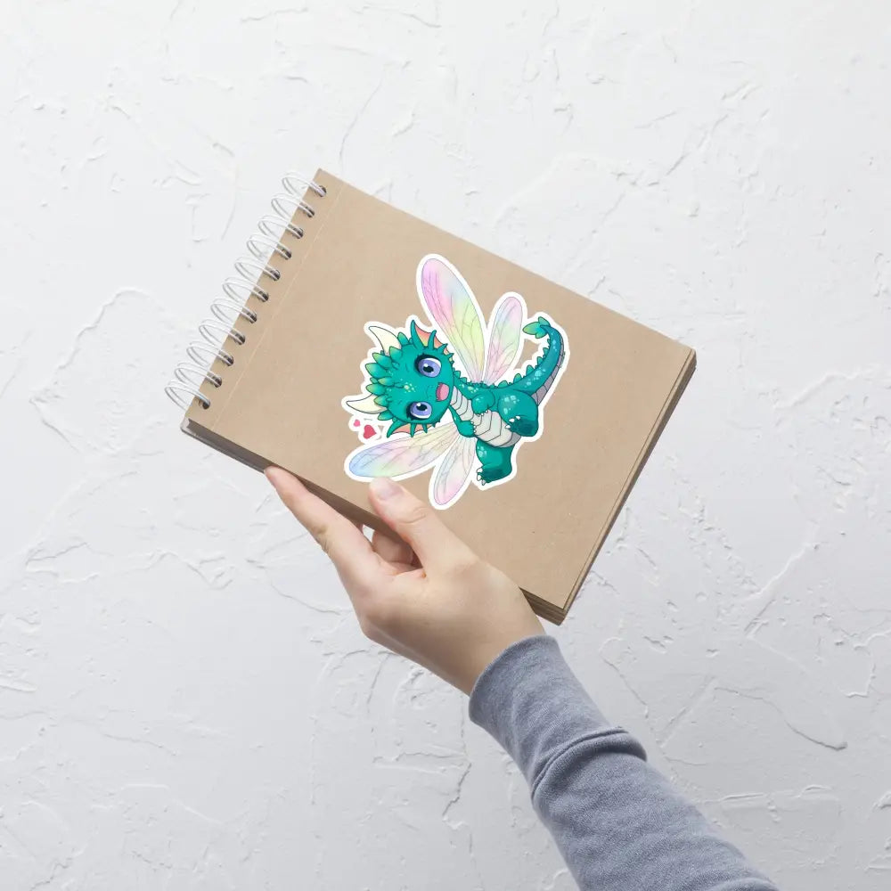 A product mockup of a 5x5" vinyl sticker on a brown notebook cover, featuring Tetra the Fairy Dragon.