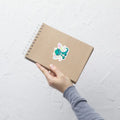 A product mockup of a 3x3" vinyl sticker on a brown notebook cover, featuring Tetra the Fairy Dragon.