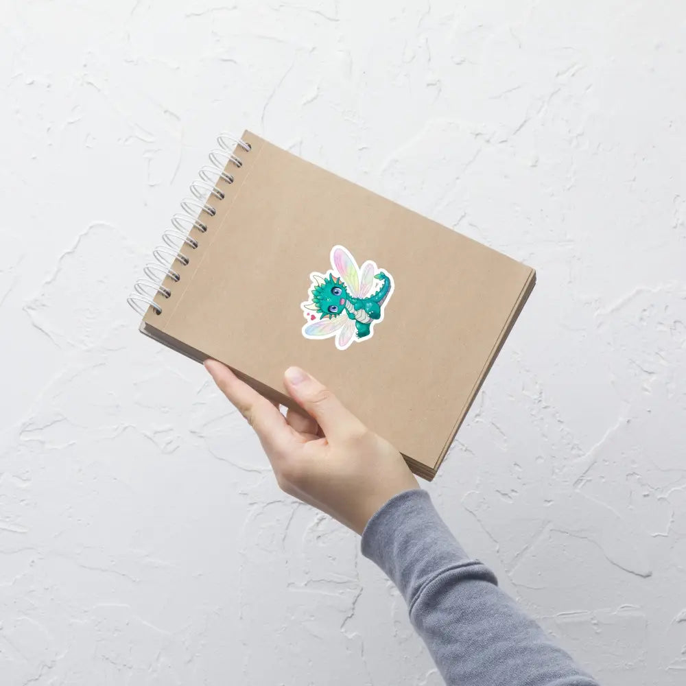 A product mockup of a 3x3" vinyl sticker on a brown notebook cover, featuring Tetra the Fairy Dragon.