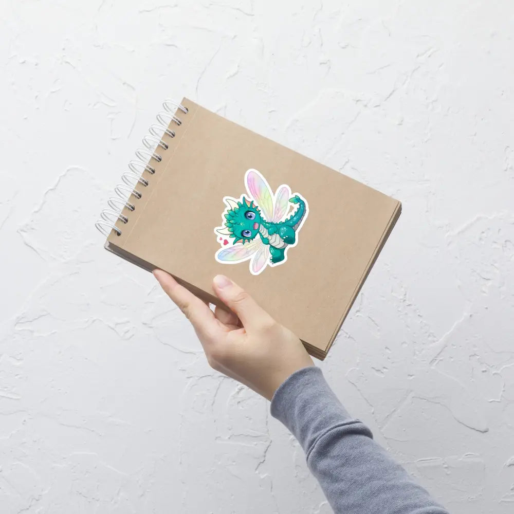 A product mockup of a 4x4" vinyl sticker on a brown notebook cover, featuring Tetra the Fairy Dragon.