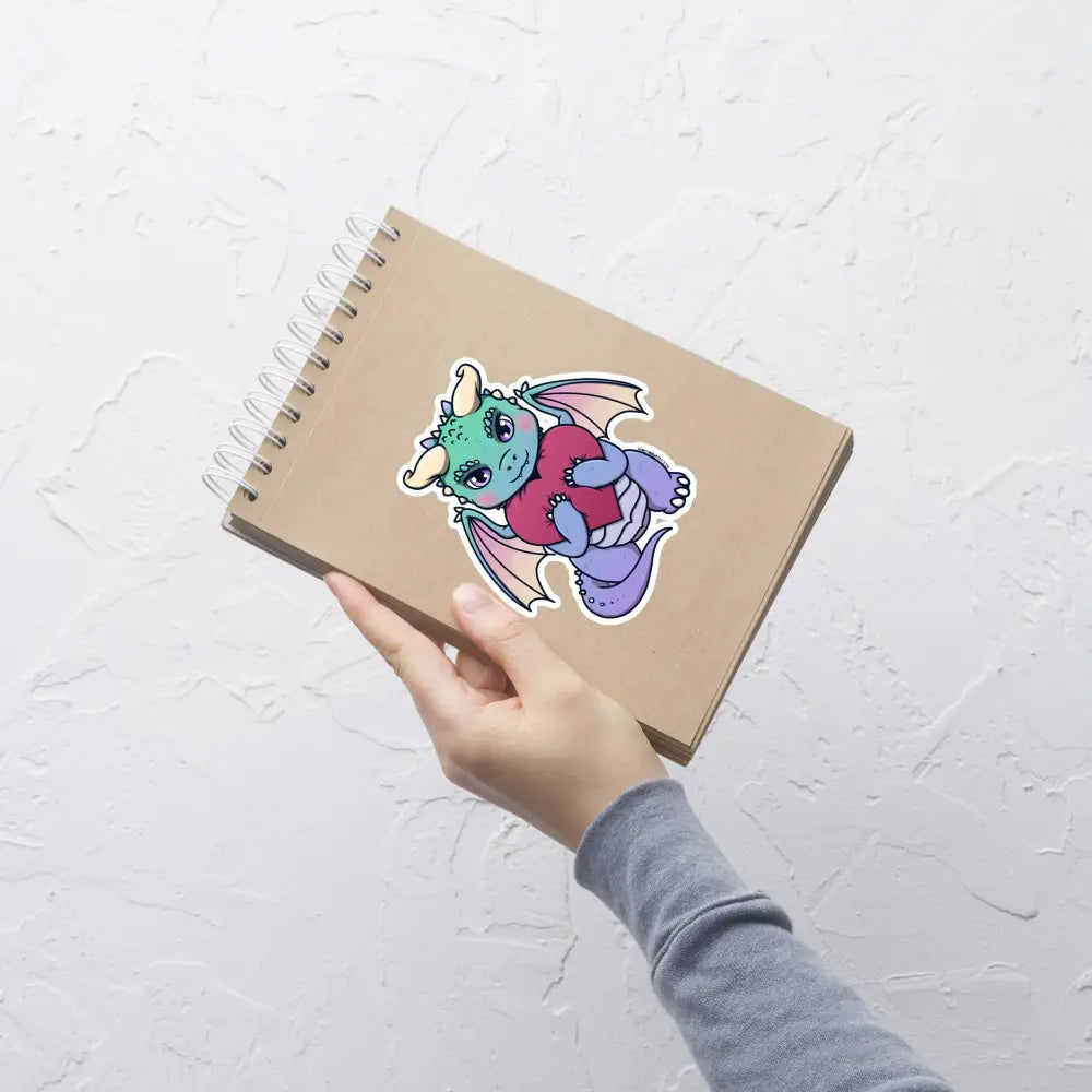 A product mockup of a 5x5" vinyl sticker on a brown notebook cover, featuring Vivian the Dragon.