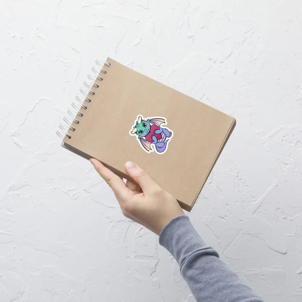 A product mockup of a 3x3" vinyl sticker on a brown notebook cover, featuring Vivian the Dragon.